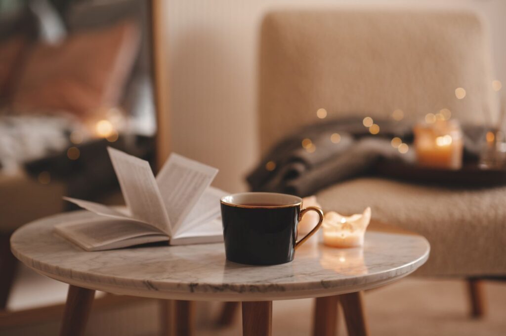 cup of tea with paper open book and burning scented candles on marble table over cosy chair and glowing lights in bedroom closeup