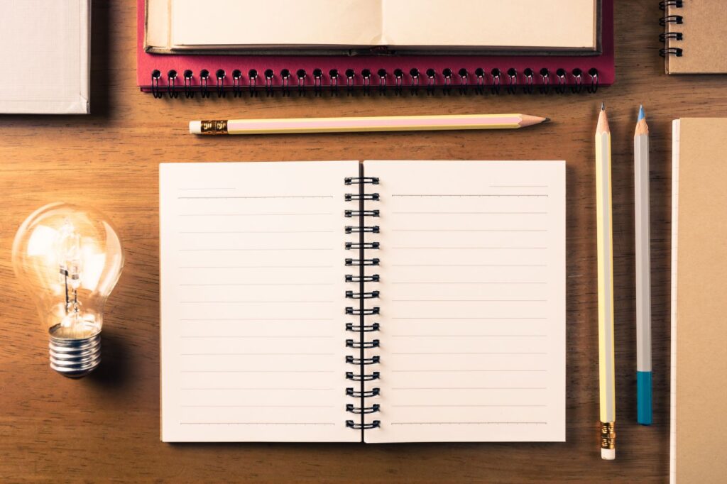 An overhead shot of an open, blank spiral notebook centred on a wooden desk, surrounded by pencils, a glowing light bulb, and other stationery.