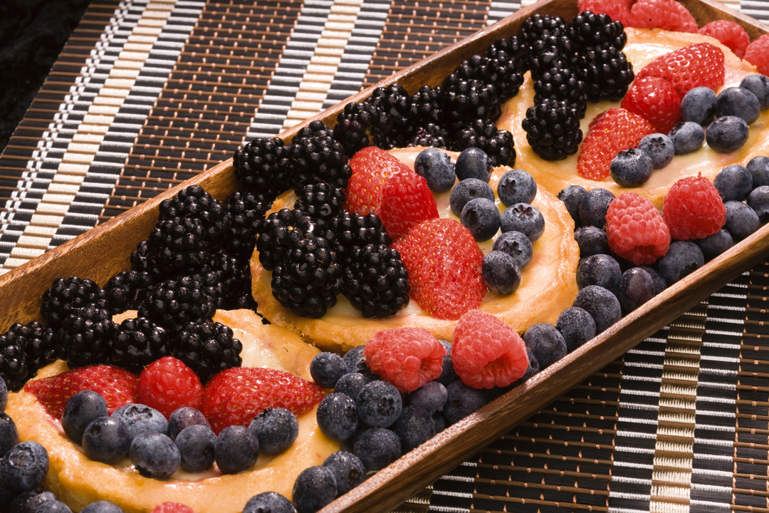 A long, wooden serving tray holding several round custard tarts topped with an abundance of fresh blackberries, blueberries, strawberries, and raspberries.