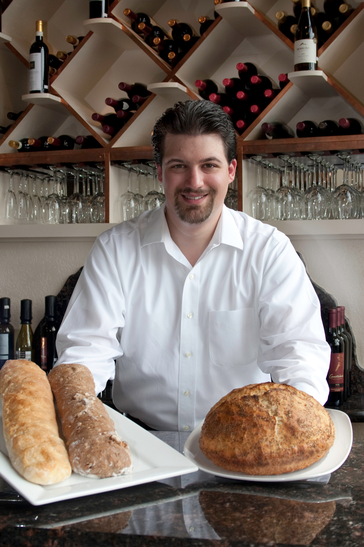 A man in a white collared shirt stands behind a counter holding two plates of artisanal bread
